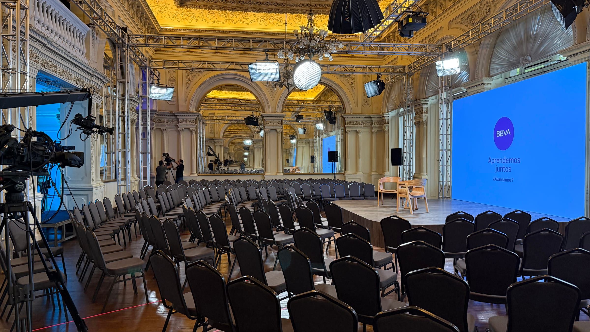 Salón con escenario, pantalla LED con el logo de BBVA y sillas, con cámaras de transmisión sobre trípodes
