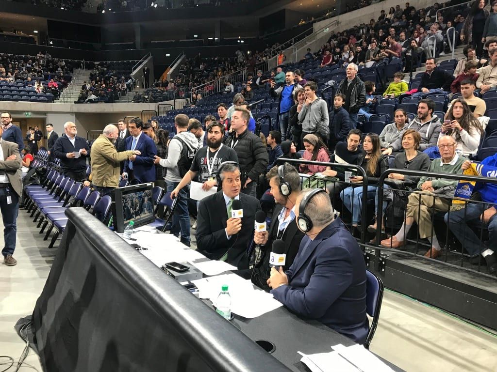 Comentaristas con auriculares y micrófonos en la mesa de transmisión junto a la cancha, con el público detrás