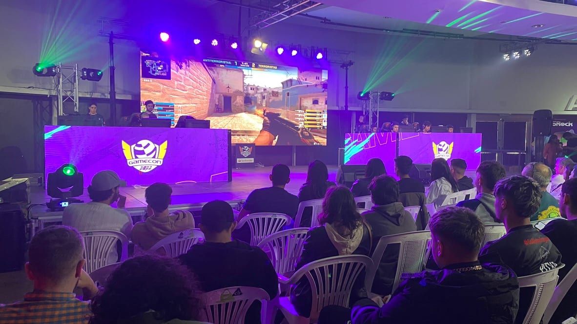 Público sentado frente al escenario de Gamecon 2022 con el logo del evento y una partida proyectada en pantalla