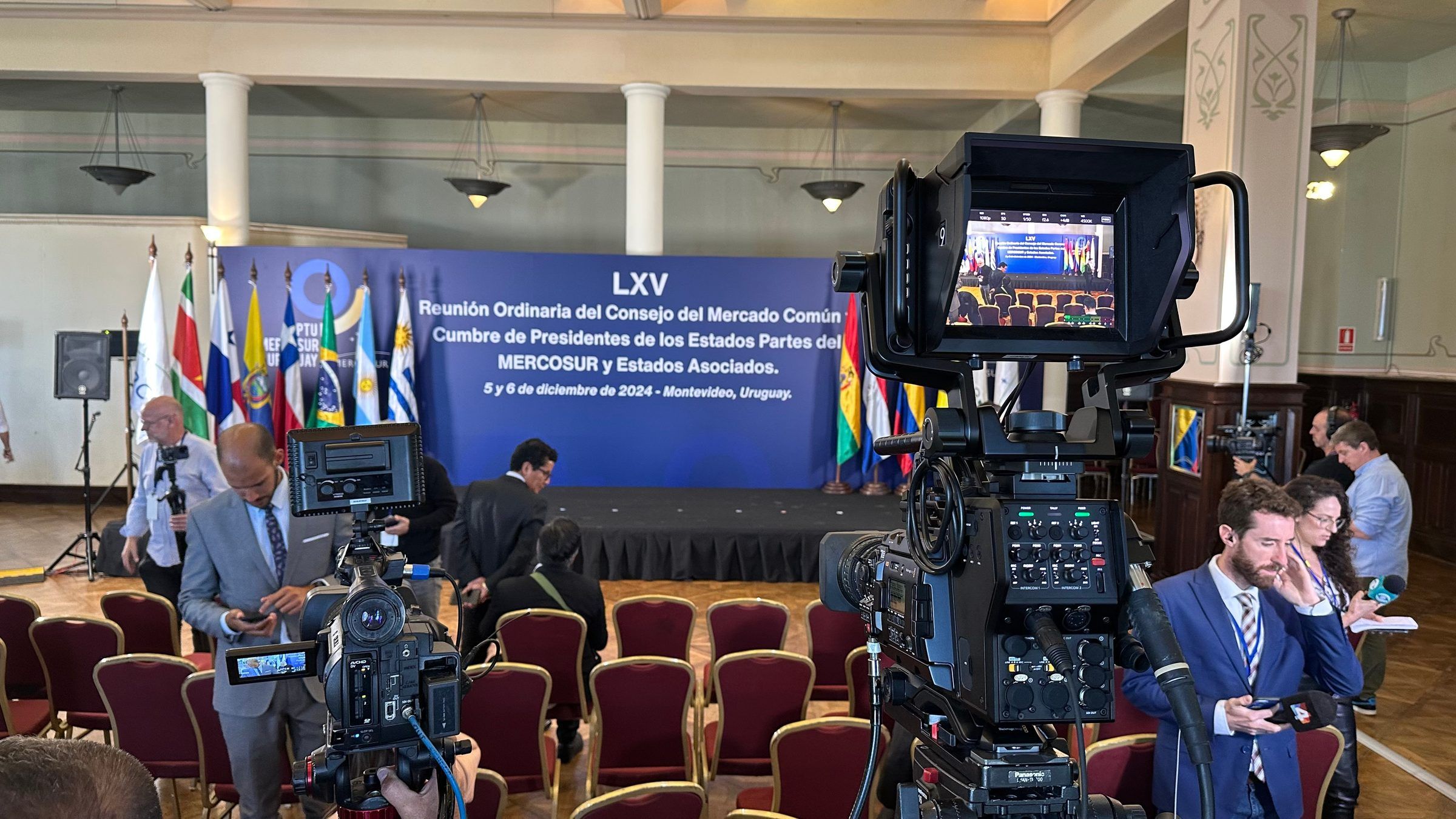 Cámara de transmisión en primer plano frente al cartel de la LXV reunión del Consejo del Mercado Común, en Montevideo.