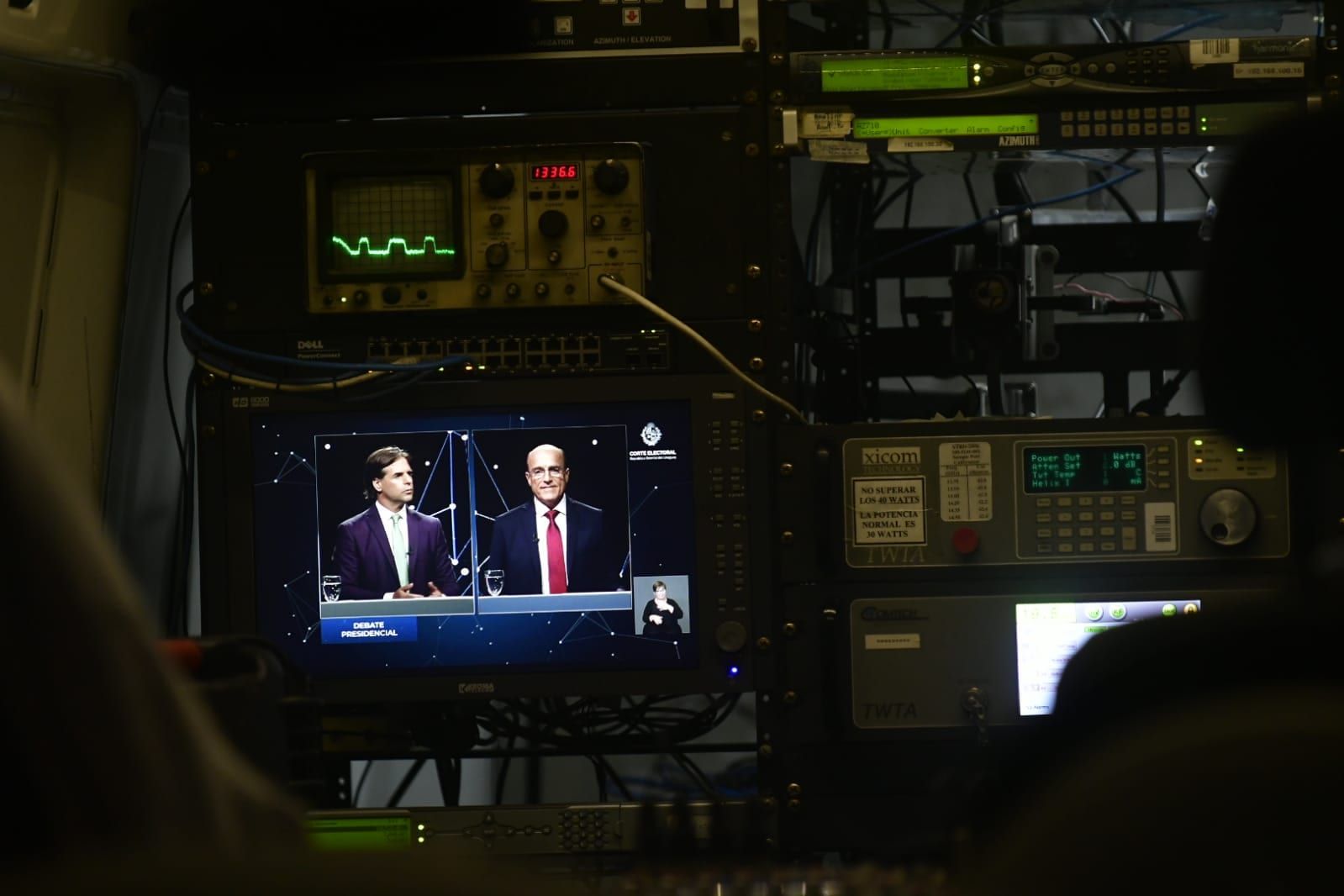 Equipos técnicos de transmisión con un monitor que muestra la señal del debate, con los dos candidatos en pantalla dividida y el logo de la Corte Electoral