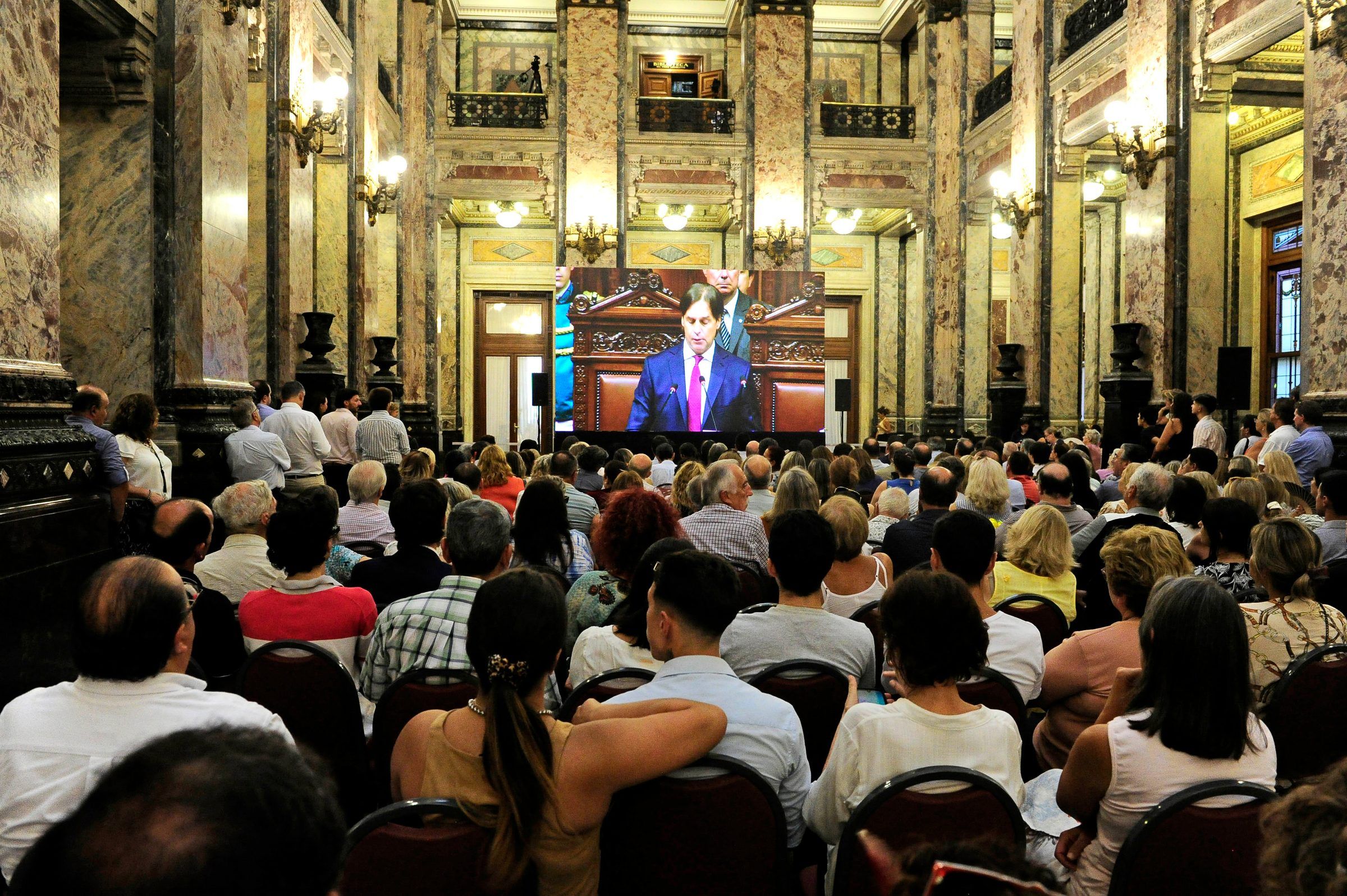 Público sentado en el hall del Palacio Legislativo siguiendo el mensaje por una pantalla.