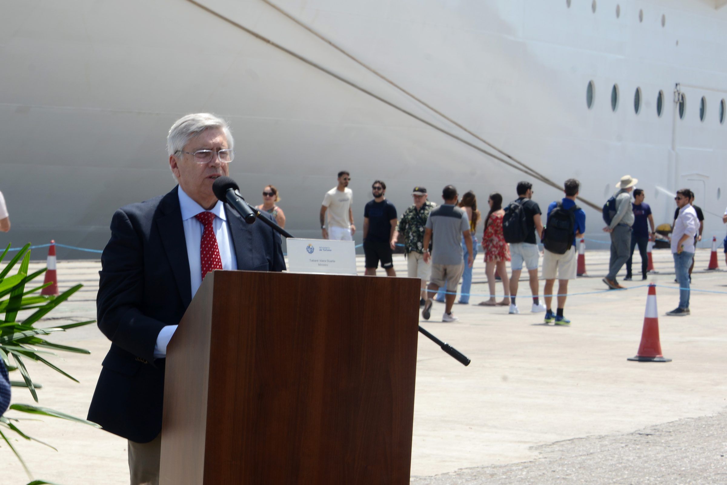 Discurso en el atril de la ceremonia de lanzamiento, con el casco del crucero y turistas al fondo.