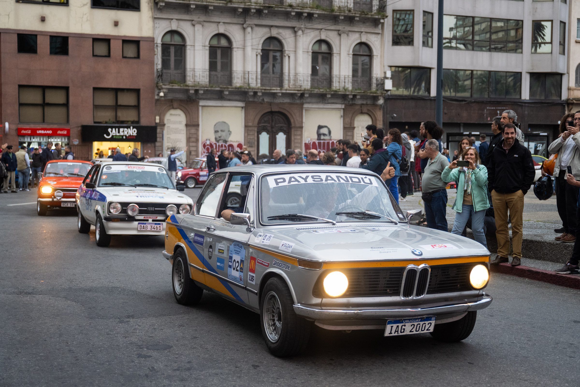 BMW 2002 de época avanza con los faros encendidos entre el público durante la largada simbólica