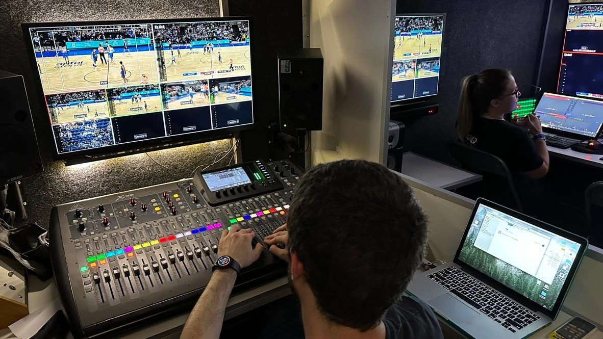Operador de audio en la consola de la unidad móvil, con monitores que muestran los partidos y una integrante del equipo al fondo
