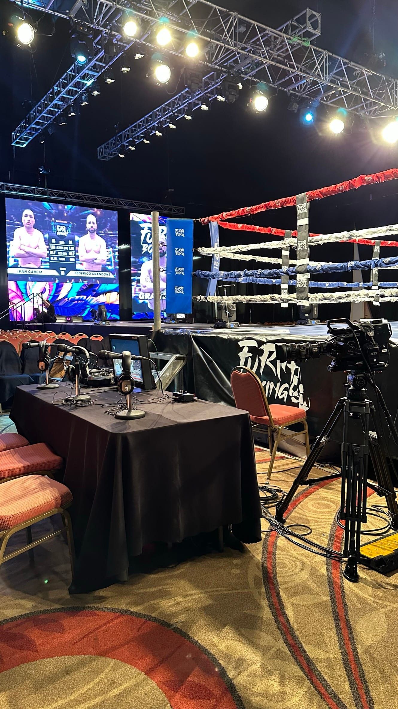 Cámara sobre trípode junto a la mesa de comentaristas y el ring, con las pantallas led al fondo